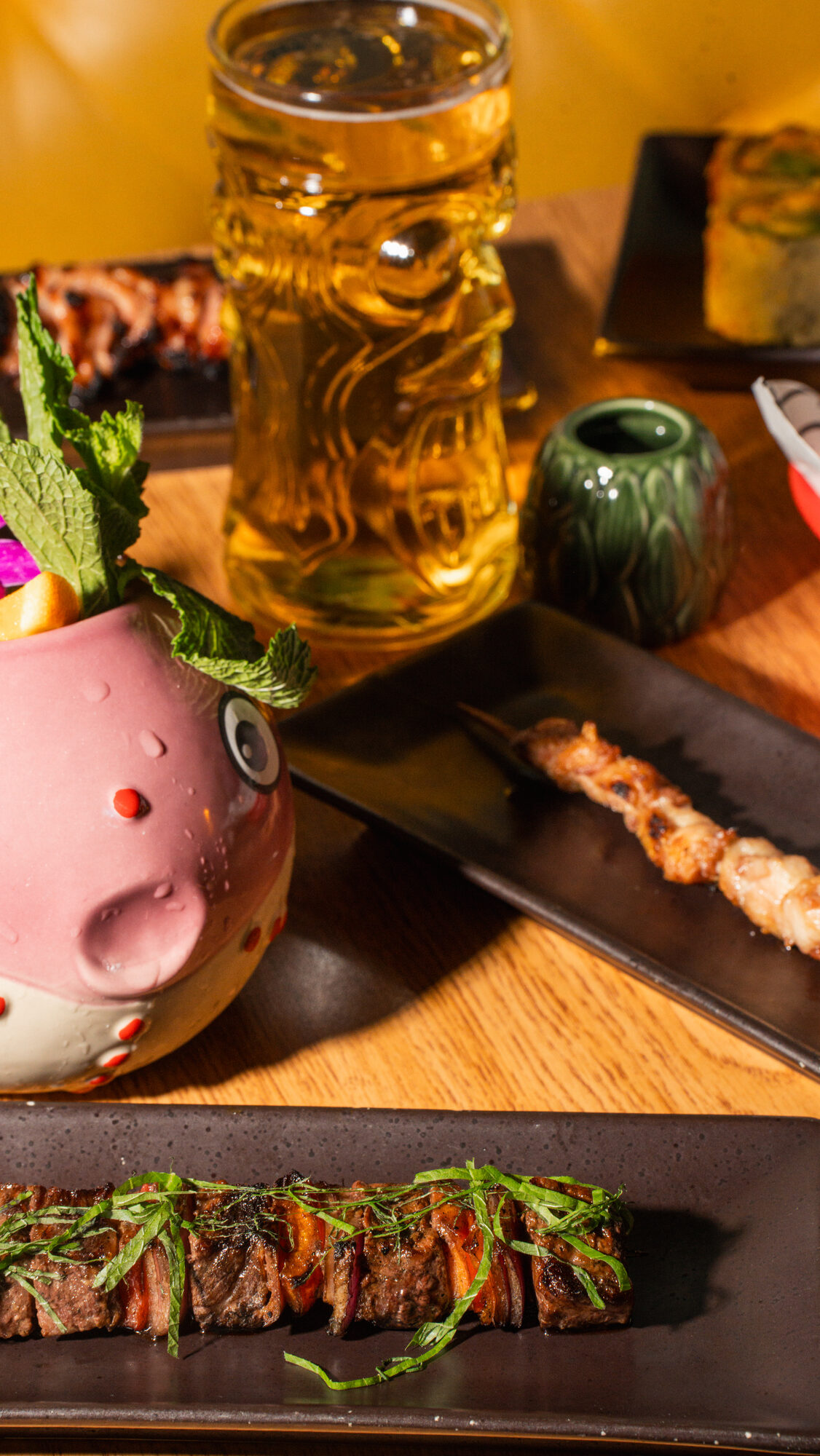 Tiki mug, a stein of beer, robata skewers and steak on a Hojoko table
