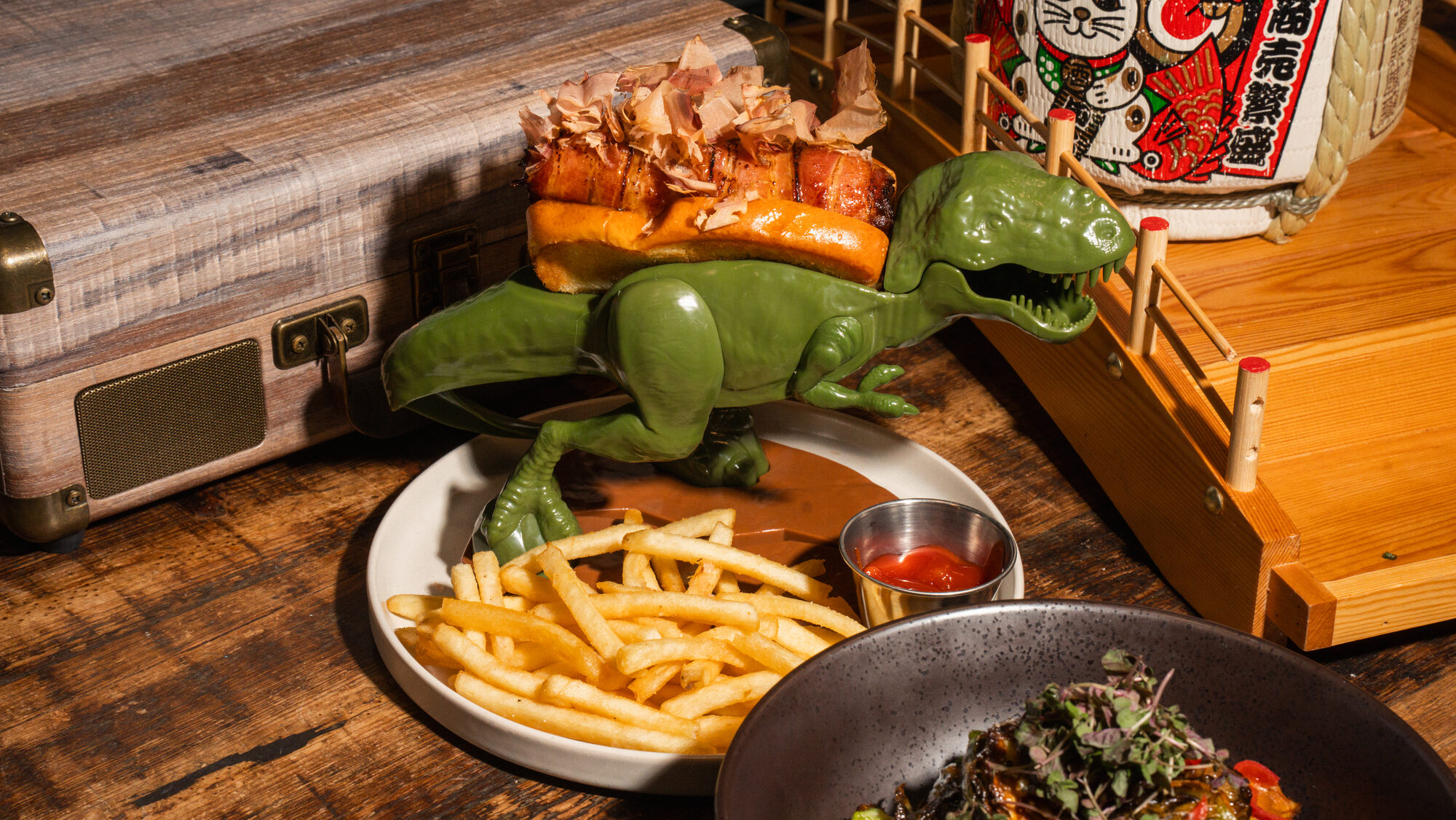 Doggzilla hot dog held by a toy dinosaur, with fries and sake